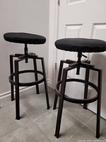 Two black metal frame bar stools with round brown wood veneer seats and black cushions. Shown from front angle against wall.
