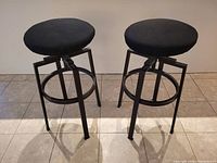 Set of two bar stools with round brown wood veneer seats and black removable cushions. Black metal legs with height adjustment mechanism and circular footrests.