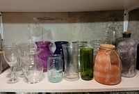 Shelf holding 5 clear twisted stem wine glasses, a selection of clear, green, purple, and blue glass vases, and two larger clay-look decorative glass items with cork finishes.