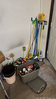 Photo showing various brooms, mops, and plastic buckets arranged against a wall next to a door.