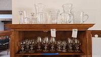 Full view of pressed glass vases and pitchers on china cabinet top with glass goblets below.