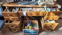 Wide view of desk with multiple open drawers revealing various office supplies including pens, markers, highlighters, paper clips, and small boxed envelopes.