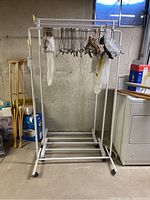 Full view of one white tubular metal rolling clothes rack with baby hangers attached, positioned against a concrete wall in a basement.