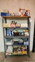 Full shelving unit with multiple boxes and supplies including books, threads, paints, buttons, and other items on metal shelving in basement.