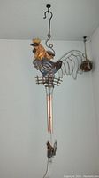 Metal rooster windchime hanging on wall, showing multicolored metal rooster on small fence with four copper-tone chimes below and metal spherical potpourri holder hanging nearby.