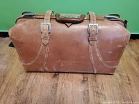 Front view of brown leather suitcase showing buckle straps and leather surface with wear marks.