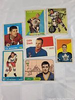 Seven vintage hockey cards laid out on a white surface, showing players in various poses wearing historic NHL team uniforms from 1950s to 60s.