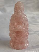 Front view of the carved pink quartz Buddha figure, showing detailed facial features, hair, robe folds, and hands resting on knees.
