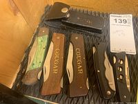 Five folding pocket knives arranged on a black tray, including two Cougar wooden handled knives, one green handled knife, two black handled knives, and a leather sheath.