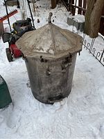 Side view of metal distiller showing cylindrical body with weathered surface and octagonal lid with central vertical pipe handle.