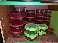Multiple round glass food storage containers with red lids and some with green lids arranged on a wooden shelf.
