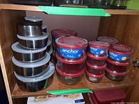 Stacked stainless steel containers with white plastic lids and several red-lidded Anchor glass containers inside a wooden cabinet.