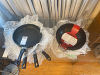 Six frying pans displayed on bubble wrap, including four T-fal frying pans and two Cuisinart woks.