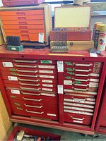 Front view of red metal cabinet showing two columns of drawers labeled for screws, drill bits and other hardware categories. Top holds drill bit cases and cans.
