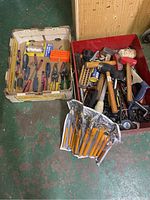 Photo of two boxes full of assorted hand tools including mallets, screwdrivers, and various small tools with two zip-up plastic cases holding jumbo punch and chisel sets in orange handles placed in front.