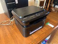 Side angle showing Samsung multifunction printer on wooden surface with power cables visible.