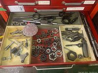 Wide view of drawer containing the main assortment of taps, dies, grips, and thread gauge disc. Various metal threading tool components are arranged, showing a typical mix of sizes and shapes.