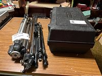 Three tripods (two lightweight aluminum, one heavy-duty steel) next to a Bausch & Lomb hard case
