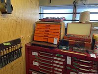 Wide view of orange tool box with small drawers, drill bit cases, chisels mounted on wall, and red metal drawer units with labeled tools
