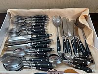 Top view of full utensil set including forks, knives, spoons, and serving spoons all with black handles and metal rivets arranged in cream fabric-lined box.