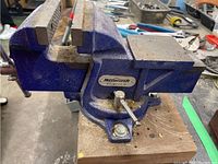 Side view of blue Mastercraft bench vise mounted on wooden work table, showing overall condition and mounting bolts