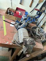 Photo showing bench vise mounted on worktable, several blue-handled clamps, bowl of nuts and bolts, and a box with hardware parts.