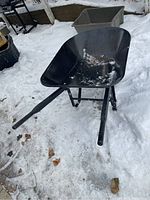Black metal wheelbarrow sitting in snow, showing tray shape, handles, and paint wear.