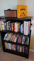 Front view of the black wooden bookcase filled with assorted books and some items on top including a brown woven basket and yellow Gobblet! box