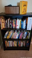 Full view of black bookshelf with three shelves filled with a variety of books, and storage basket and box on top.