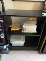 Front view of black laminate cabinet with items inside showing the fixed shelf and wheels.