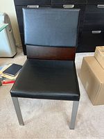 Full view of the chair showing black leather-like seat and backrest, wood back panel, and metal legs.