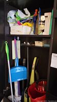Black cabinet with shelves holding various cleaning supplies, brooms, dustpan, mop bucket, and Kenmore vacuum cleaner