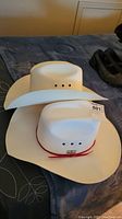 Two white cowboy hats stacked on each other showing wide brims and crown vent holes, with one featuring a red cord hatband and Calgary Stampede tag.