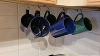 Six ceramic coffee mugs hanging from kitchen hooks showing colors and condition.