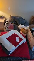 View of single bed frame with mattress, decorative pillows, and folded red and white blanket on top. Bed positioned near a wall and small shelving unit on the side.