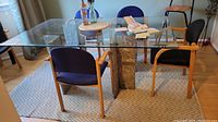 Glass top dining table with two rectangular geometrical stone supports, surrounded by chairs, placed on a patterned rug.