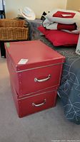 Two red metal trunks stacked on a carpeted floor next to a bed, showing metal handles and reinforced edges.