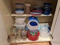 Cabinet shelf with assorted plastic Tupperware containers with lids, clear mason jars with metal lids, and two decorative Christmas tins stacked below.