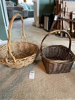 Two baskets side by side on a carpeted floor; one light round basket with twisted handle and one darker square basket with arched handle.