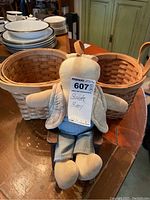 Photo showing two oval woven baskets with handles and fabric bunny doll seated in small wooden rocking chair on wooden table.