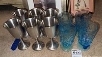 Six pewter goblets with smooth finish and pedestal base alongside four blue textured glass stem glasses with wide rims and footed bases.