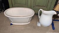 Photo of vintage white ceramic pitcher next to large oval bowl on carpeted floor in front of green cabinet.