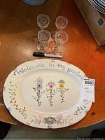 Photo of oval Pfaltzgraff ceramic plate with floral and birdhouse design and text 'Welcome to my garden', placed beside four clear cordial glasses on wooden surface with a marker for scale.
