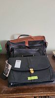 Photo of two black bags on a table: a Portfolio brand briefcase in front and an overnight bag behind with brown leather handle.