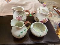 Four ceramic pieces: a Belleek creamer and sugar bowl with shamrock design, and a Brazilian creamer and lidded sugar bowl with pink flowers and handles, placed on a dark wood surface.
