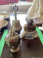Three brass decorative items on a table; includes a bell, a seated Buddha statue, and an ornate head sculpture.