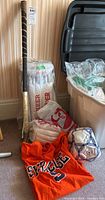 Wooden field hockey stick, cricket pads in plastic packaging, orange Syracuse T-shirt, and soccer ball packed together on carpeted floor