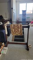 Wooden quilt rack with fabric runner and macrame hanger displayed in an attic or third story area, photo taken to show the full view of the rack and items hanging.