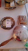 Three silver serving pieces arranged on a table: round plate, covered dish, and hinged lidded pan with wooden handle.