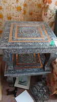 Detailed photo of one square aluminum side table showing ornate embossed surface pattern with copper inlay accents.